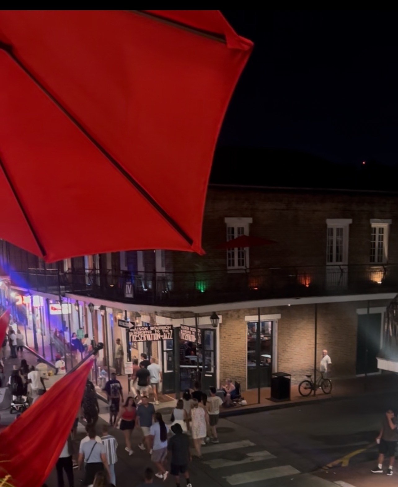 A busy nighttime street scene in Bourbon Street New Orleans with crowds walking under red umbrellas and neon-lit buildings.