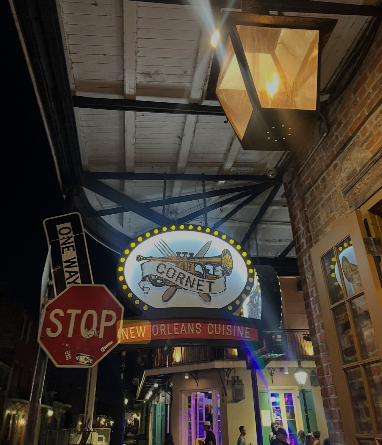 Illuminated Cornet New Orleans Cuisine sign hanging above a narrow street at night, with people below.