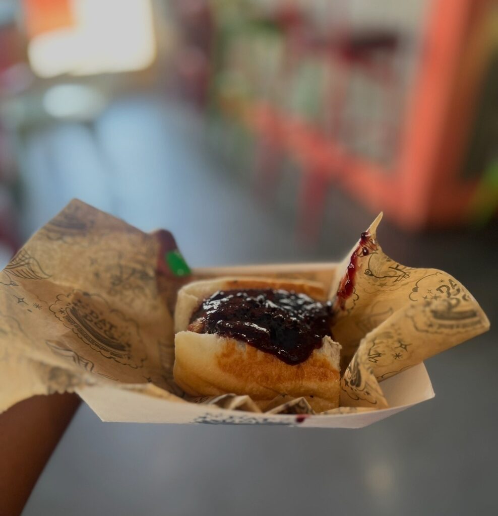 A hot dog in a bun topped with blackberry sauce wrapped in branded paper and held in a paper tray.