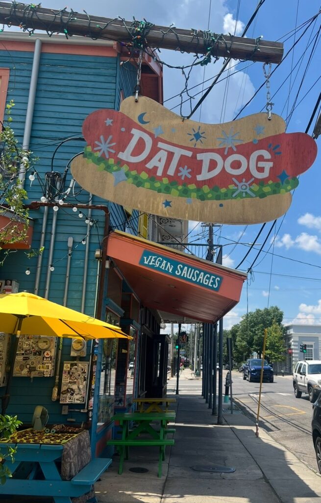 Exterior of Dat Dog restaurant with a colorful hanging sign, outdoor seating, and a sidewalk view on a sunny day. 