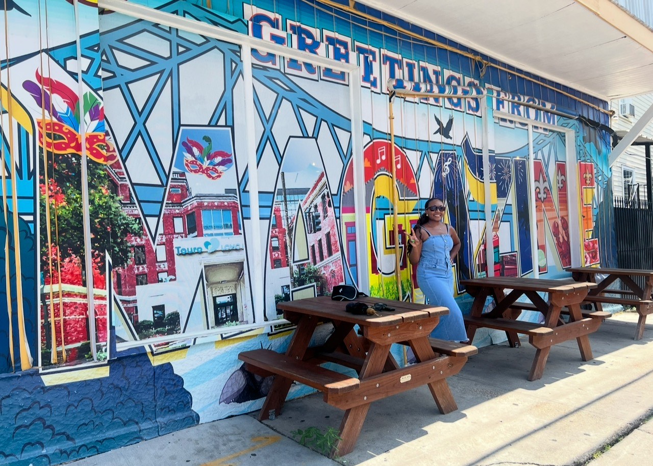 A woman posing and smiling in front of a colorful mural with picnic tables outside. 