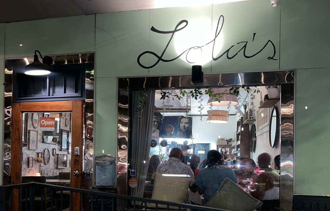 Exterior of Lola's restaurant at night, with diners visible through the window and a lit sign above the entrance. 