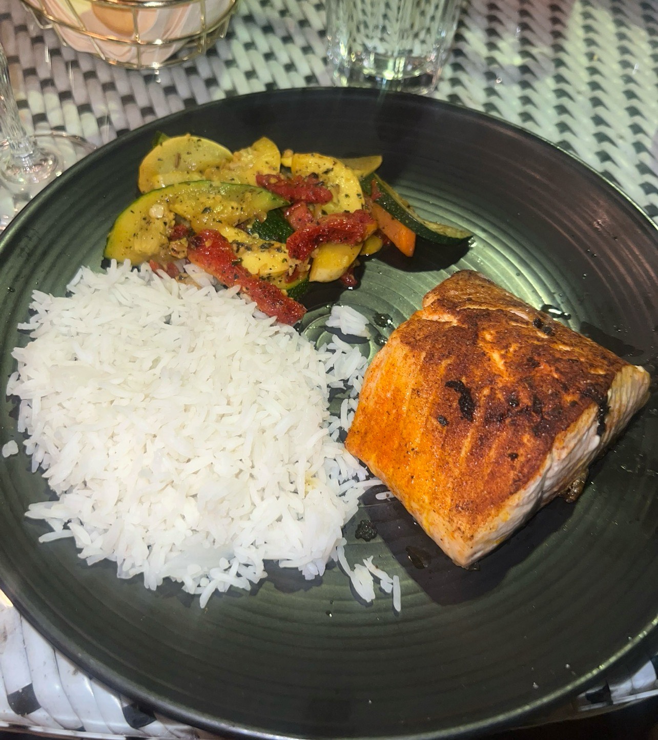 A plated meal with seasoned salmon, white rice, and sautéed zucchini and vegetables on a dark plate. 