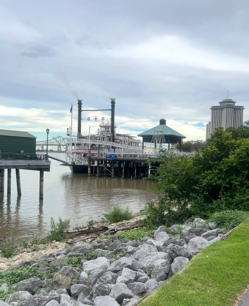A riverfront scene with a docked steamboat, pier structures and city buildings in the distance under a cloudy sky. 